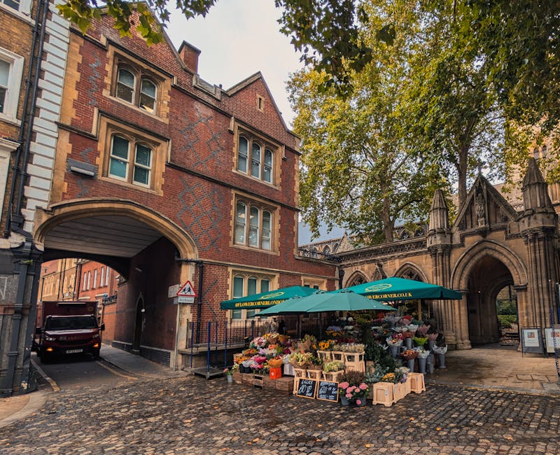 A vibrant outdoor flower stall situated beneath a green umbrella labeled 'floristconverland.com' on a cobblestone street adjacent to a historic red brick building with arched entryways. The stall disp