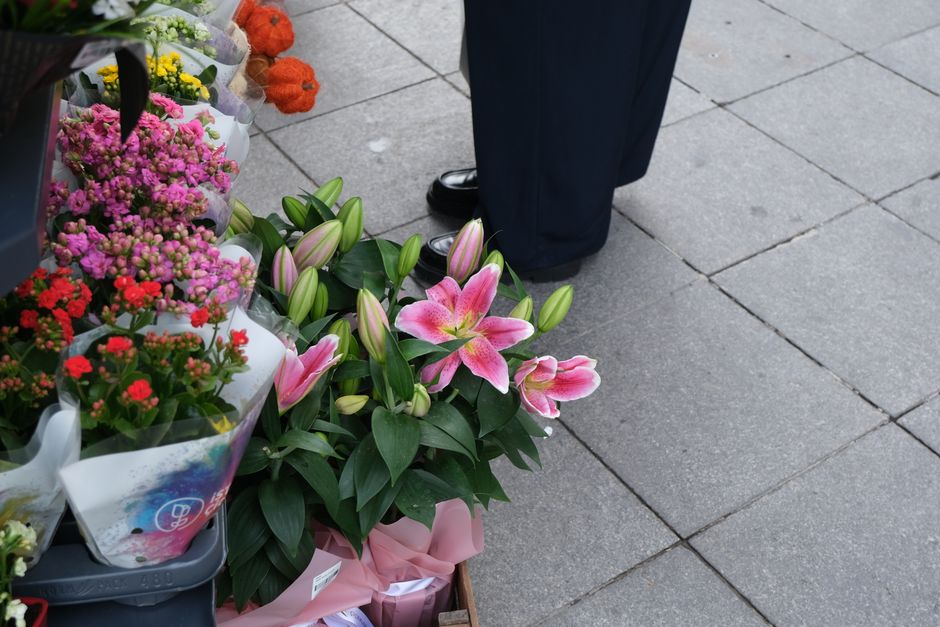 A bouquet of fresh pink lilies with large, open petals and pointed buds, arranged with glossy green leaves. The flowers are wrapped in soft pink paper, resting on a tiled sidewalk near a display of va
