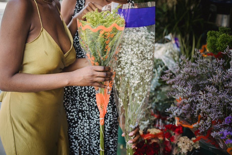 A person wearing a yellow dress holding a large, fresh bouquet of green and white flowers wrapped in clear cellophane with orange accents, surrounded by various other flower arrangements and potted pl