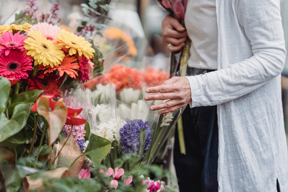 A person in a white long-sleeve top is browsing a flower stand with a variety of fresh floral arrangements, including bright pink, yellow, orange, purple, and white flowers such as daisies, hyacinths,